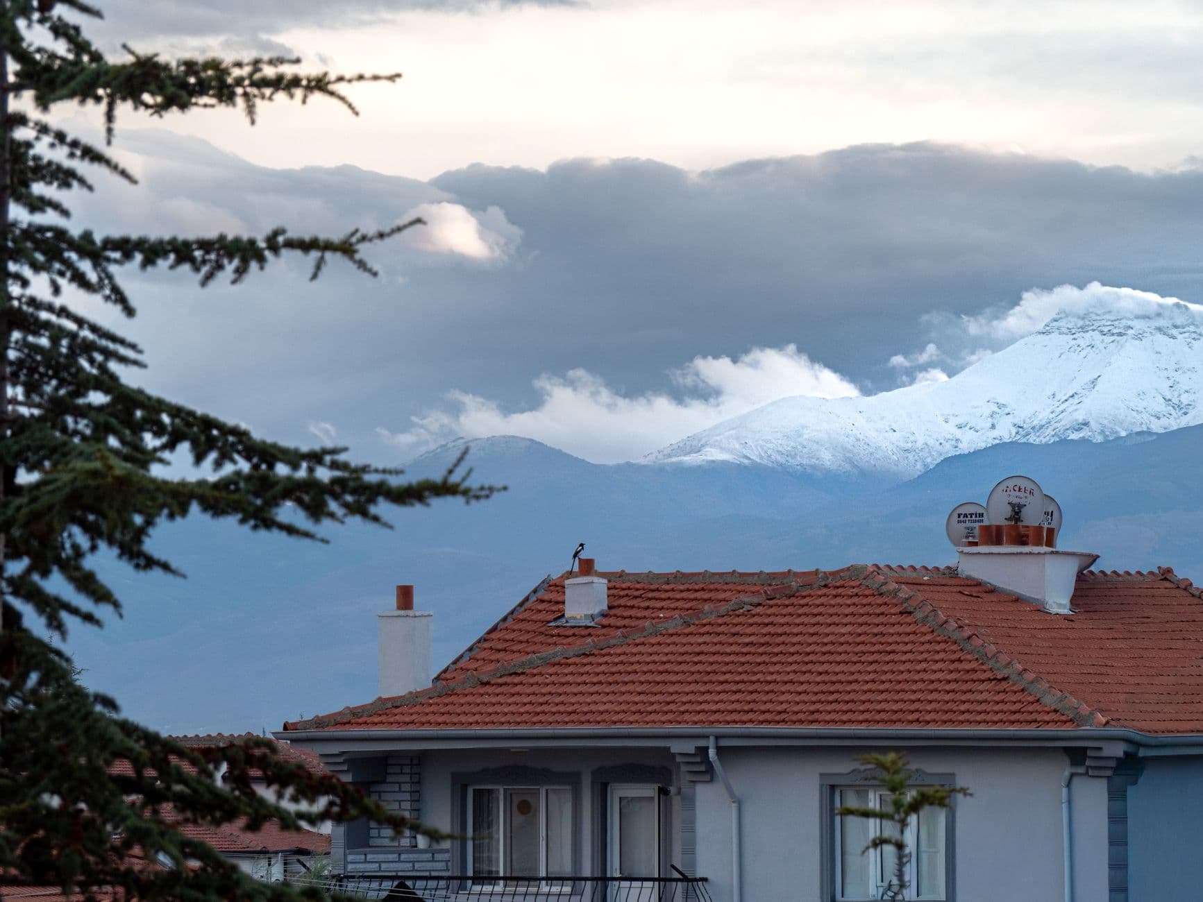 A suburban home with red roof and snow-capped mountains in the background on a cloudy day.